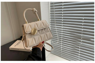 Beige quilted handbag with gold accents held by a hand, with a book and window blinds in the background.
